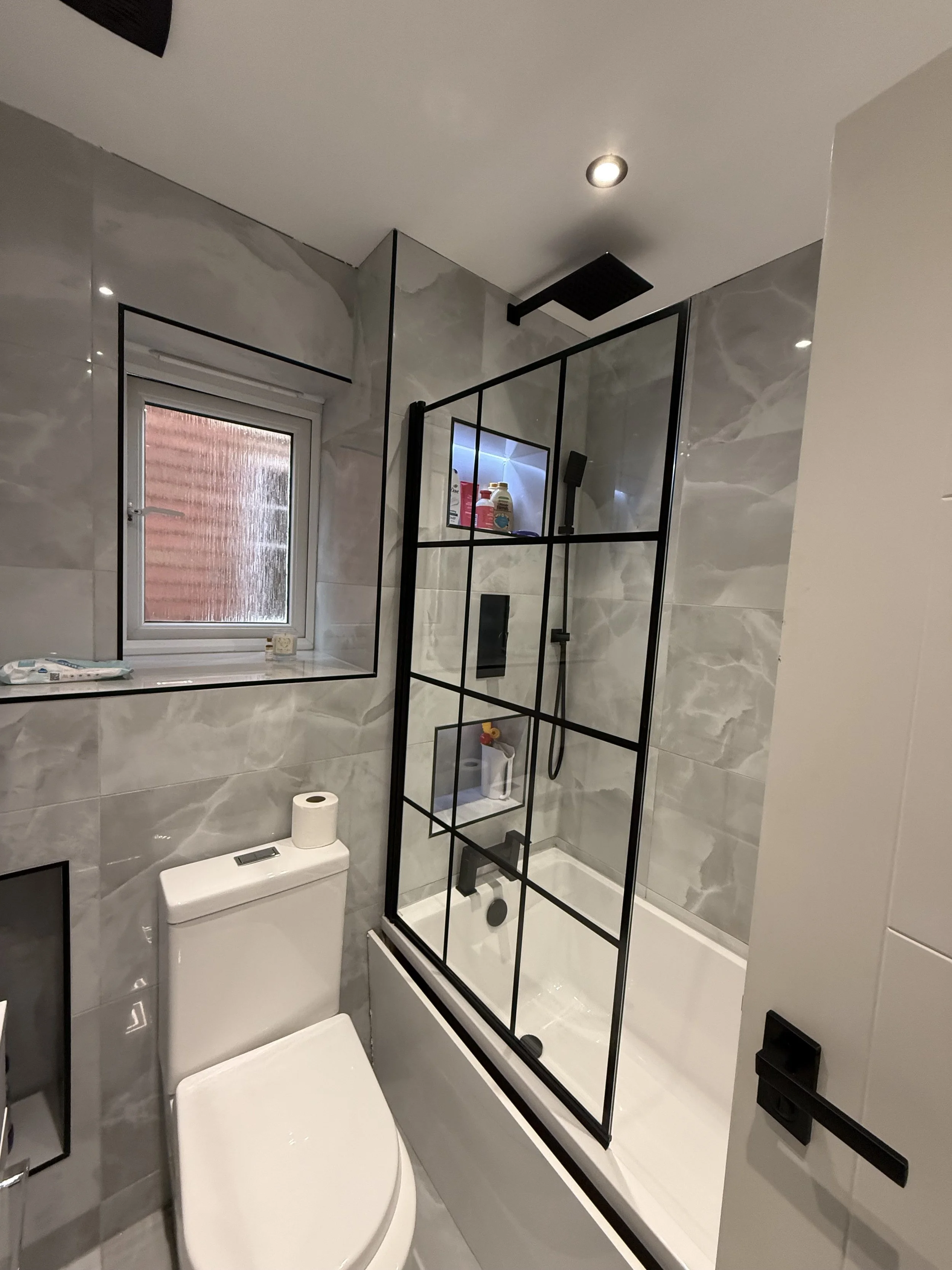A modern bathroom with gray marble walls, a small window, a white toilet, and a glass-enclosed shower with black framing.