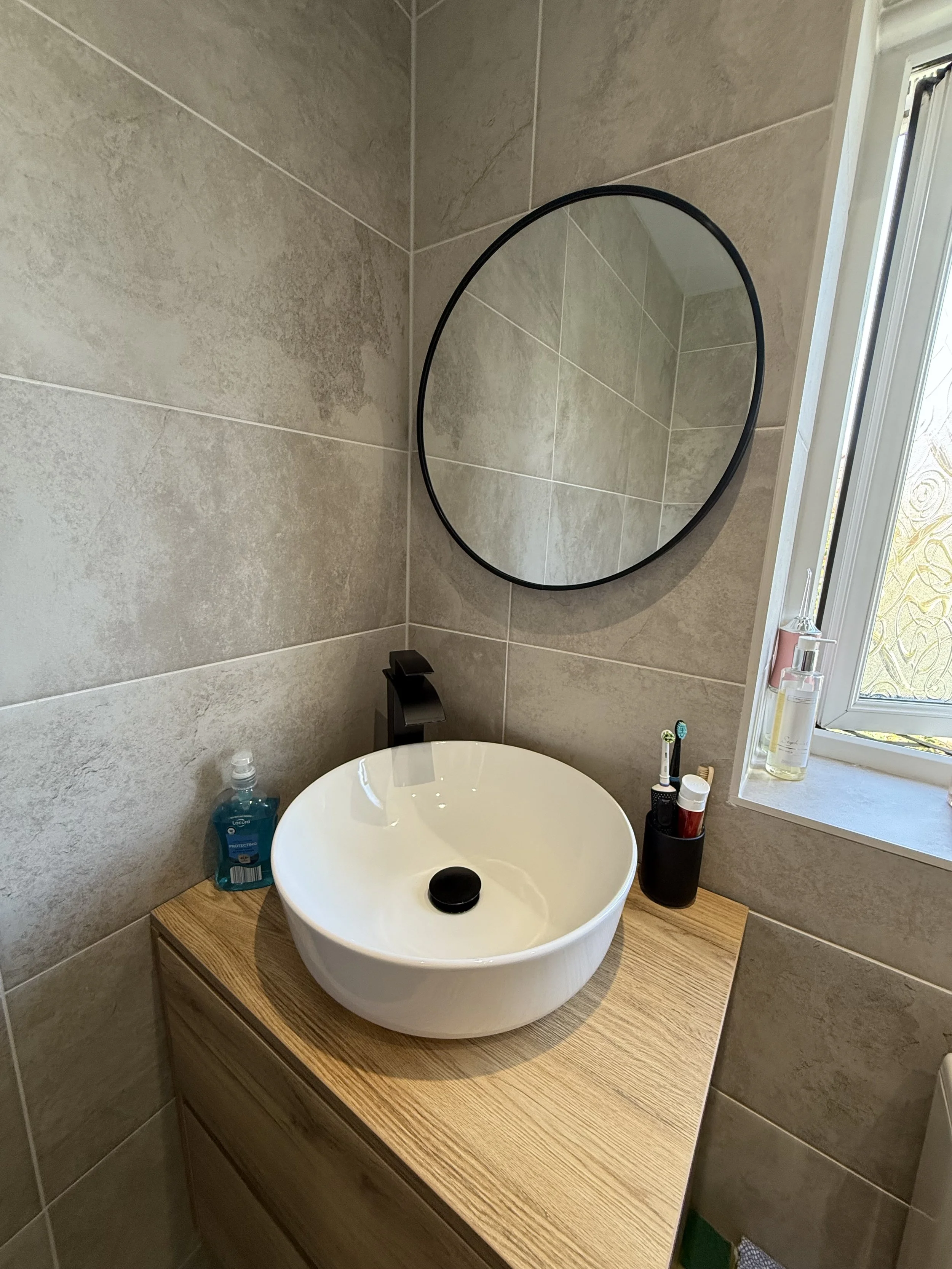 A modern bathroom with a round mirror, a white vessel sink on a wooden countertop, and toiletries including hand sanitizer, a toothbrush, and soap. There is a small window with frosted glass.