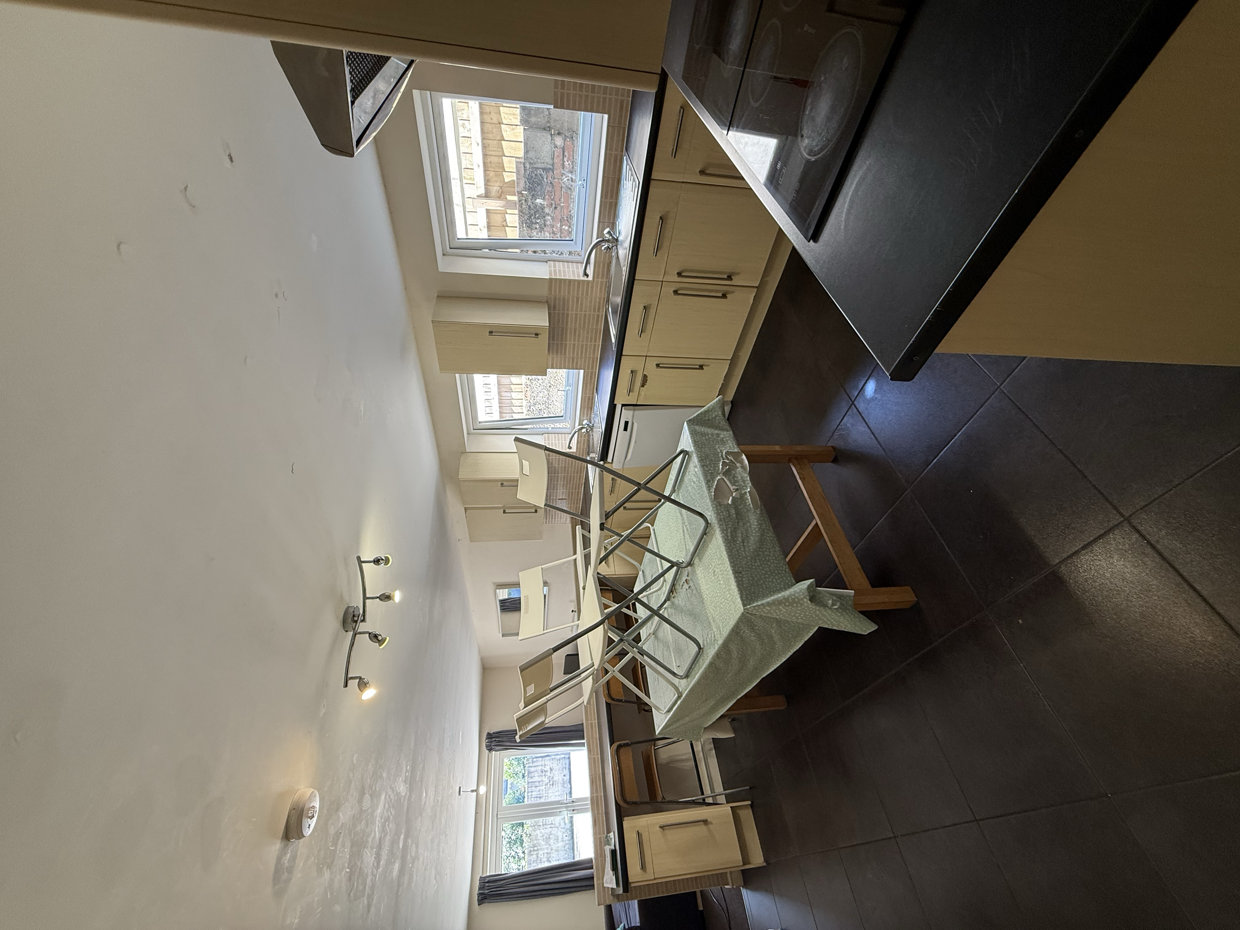 Empty kitchen with light wood cabinets, black countertop, and a table covered with a green tablecloth and a drying rack on top. There are multiple windows letting in natural light and a sliding door with dark curtains.