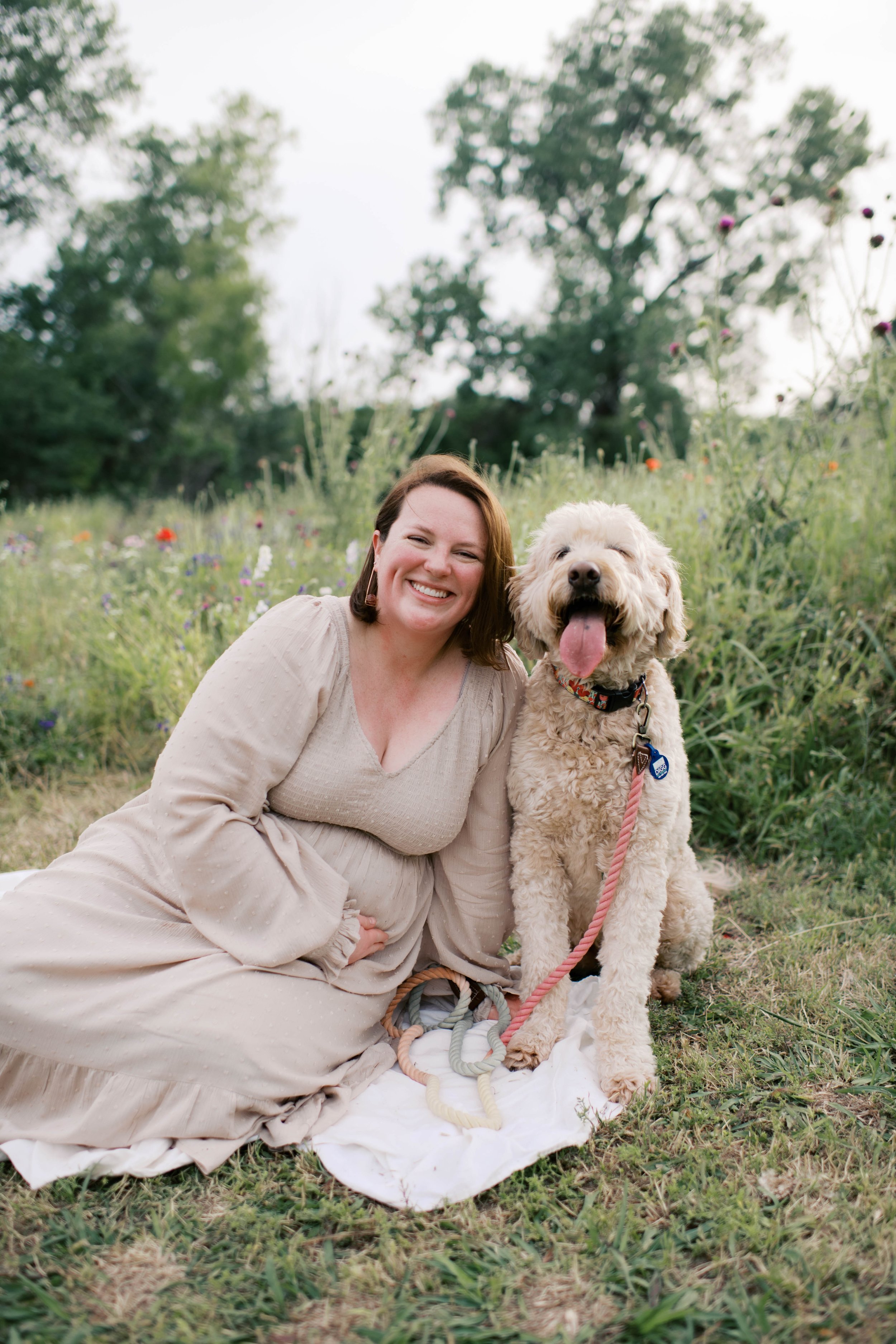A woman with dark brown hair, wearing a beige dress, sitting on a white blanket in a grassy field with flowers, smiling next to a happy, cream-colored dog with curly fur and a pink leash.
