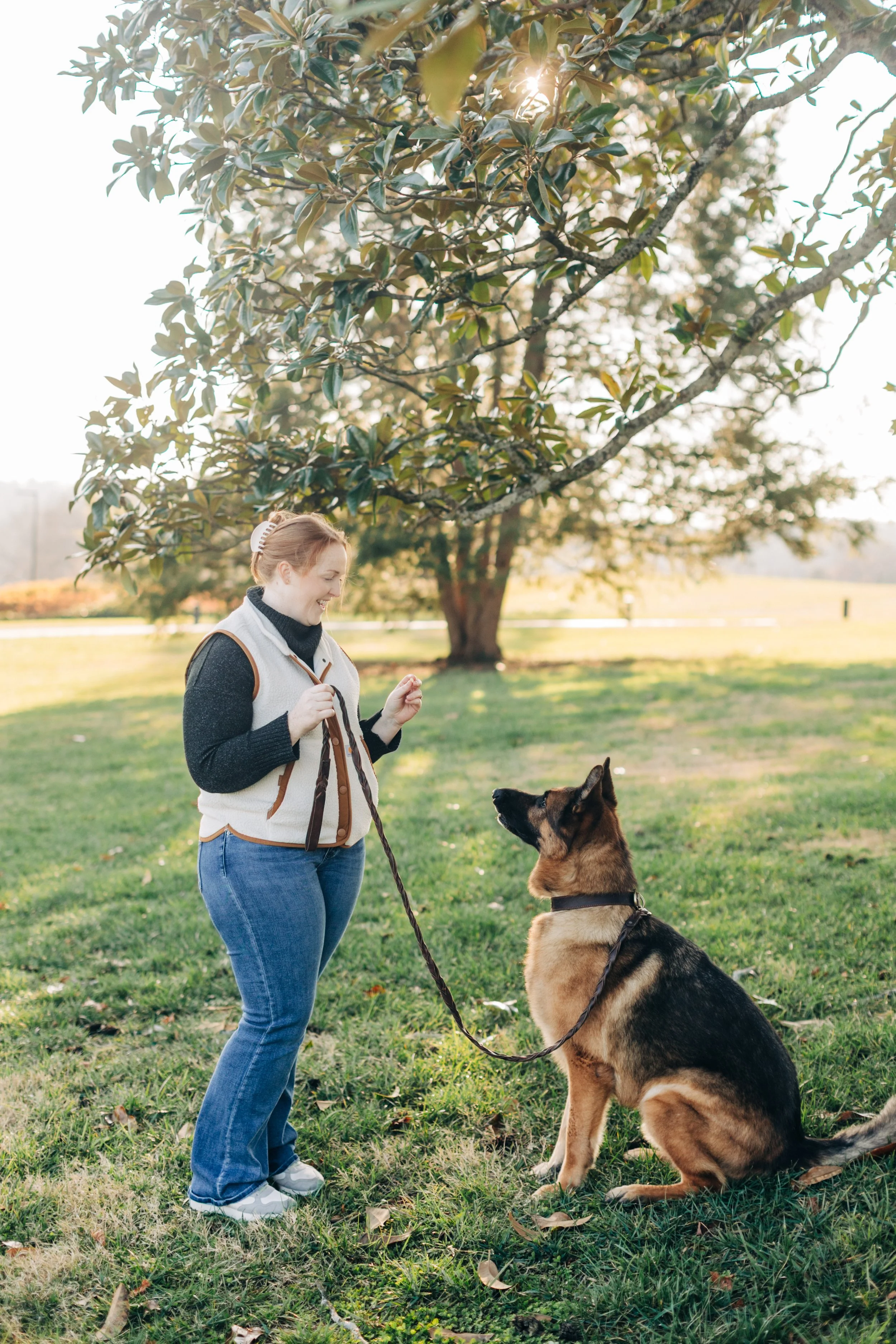 A woman with red hair, wearing a black turtleneck, white vest, and blue jeans, stands on a grassy field, holding a leash attached to a German Shepherd sitting attentively in front of her, under a large leafy tree with sunlight filtering through.