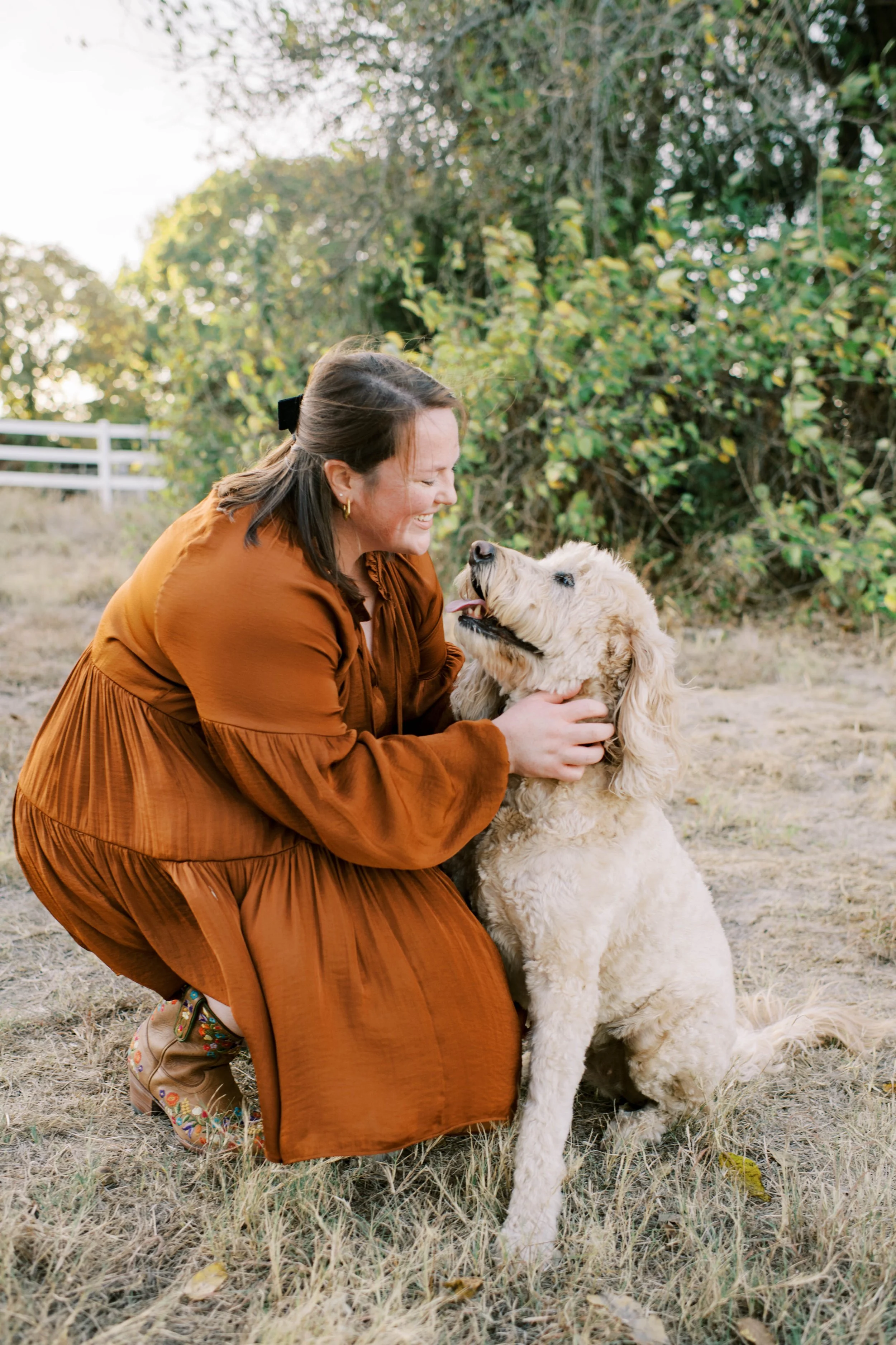 A woman with brown hair wearing a rust-colored dress smiling and playing with a golden doodle dog outdoors on a grassy field with trees and a white fence in the background.