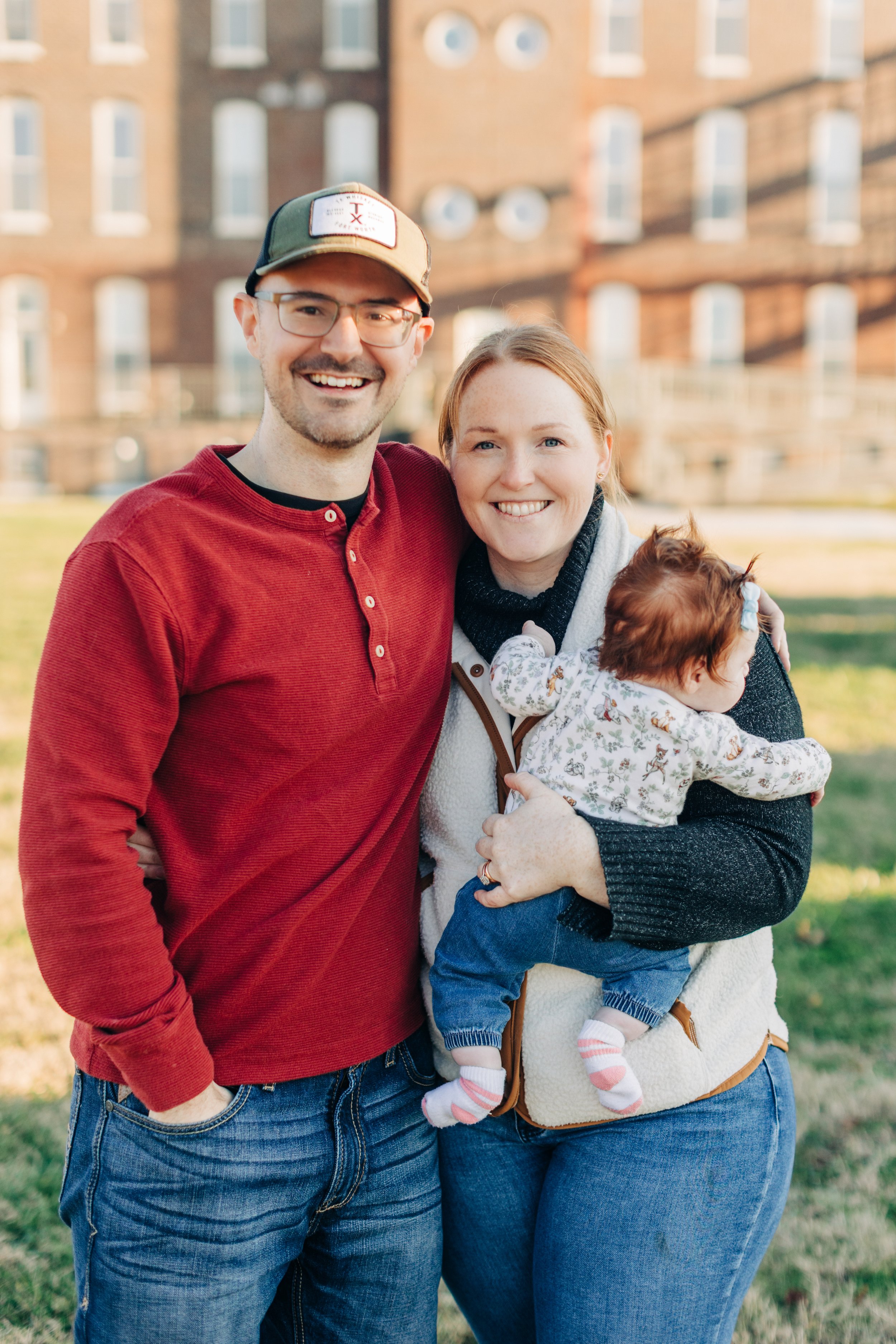 A happy family outdoors, with a man, woman, and baby girl, standing close together in front of a building, smiling at the camera.