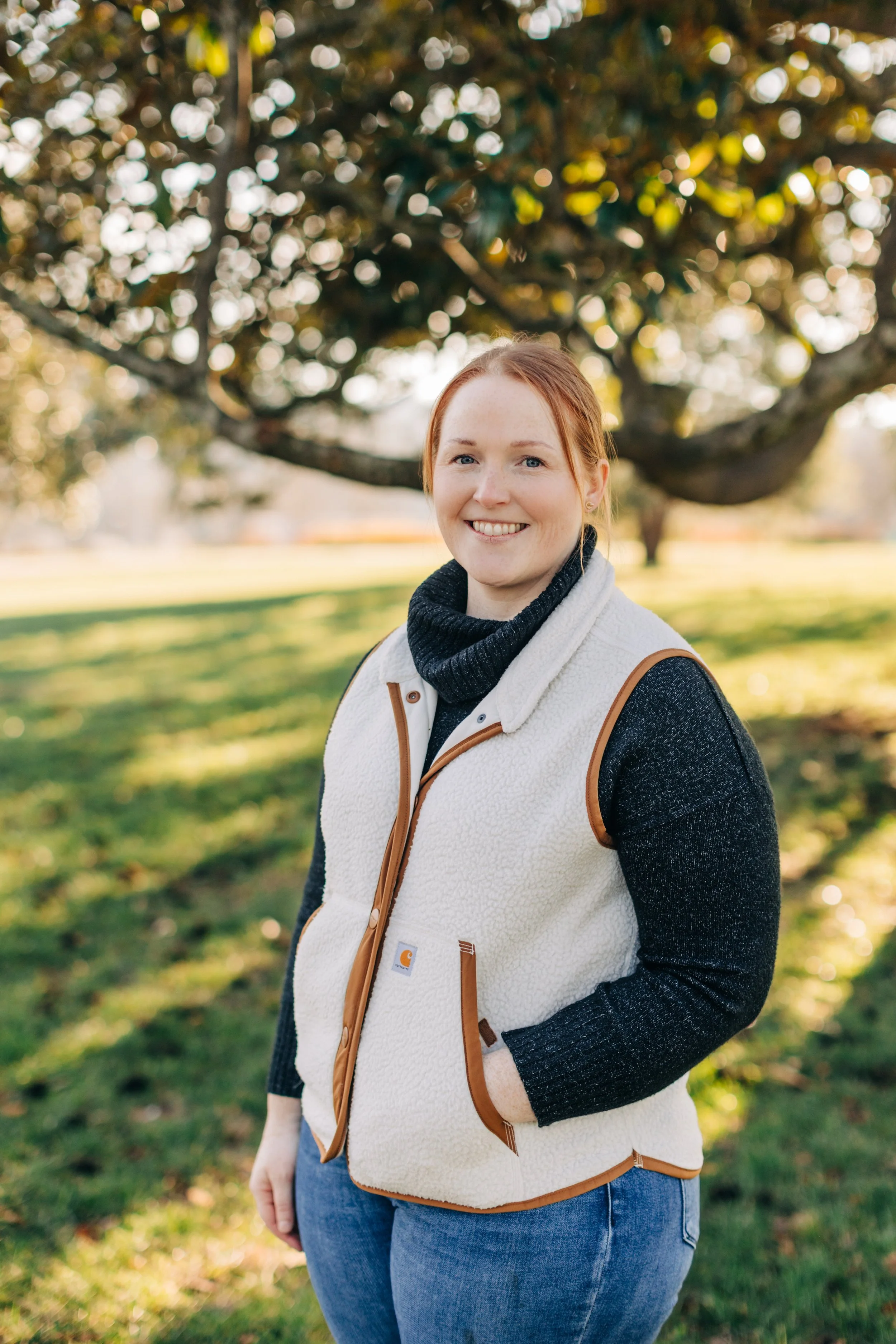 owner of Early Paws Puppy Academy, Sarah, with red hair smiling outdoors on a sunny day, standing in front of a large tree with green grass and sunlight in the background.