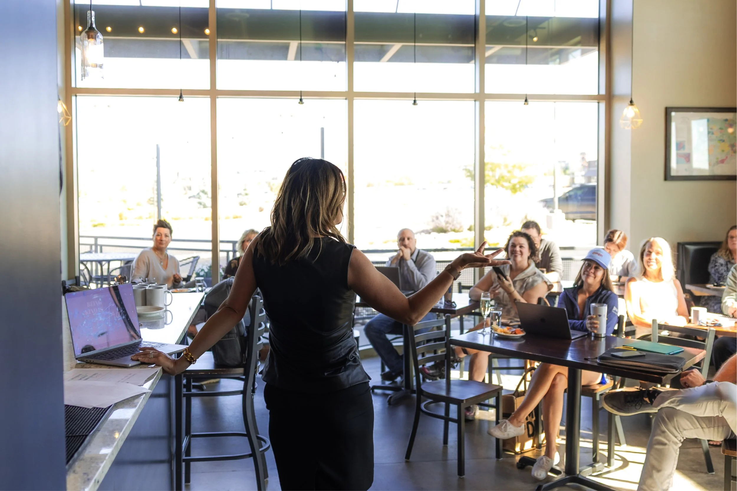 Budget with Shanon. Shanon Schinkel. A woman standing and speaking to an audience in a bright, modern cafe or restaurant. The audience is seated at tables, listening attentively. Large windows behind them let in natural light.