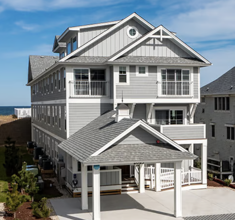 A multi-story beach house with gray siding, white trim, a covered porch, and outdoor balconies, near the ocean under a blue sky.