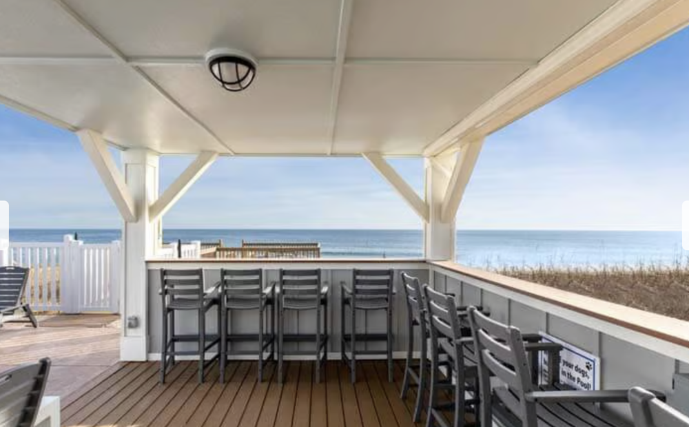 A covered outdoor deck with a row of black chairs overlooking the beach and ocean, with a blue sky and some clouds.