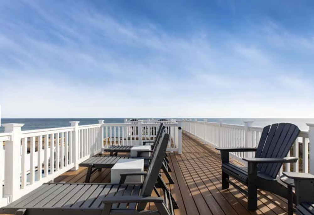 A wooden deck with Adirondack chairs and a white railing overlooking the ocean under a blue sky.