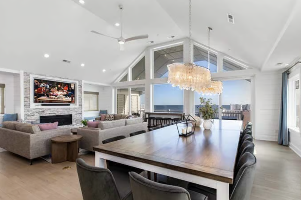 Spacious living room with high vaulted ceiling, large windows with ocean view, modern chandelier, and a wall-mounted TV above a stone fireplace. There is a large wooden dining table with black chairs and a cozy seating area with sofas and colorful cu