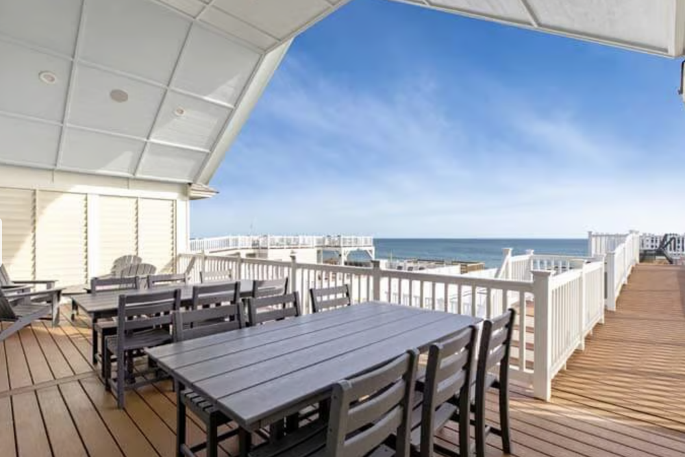 Ocean view from a deck with outdoor dining tables and chairs, white railings, and a partly roofed area.