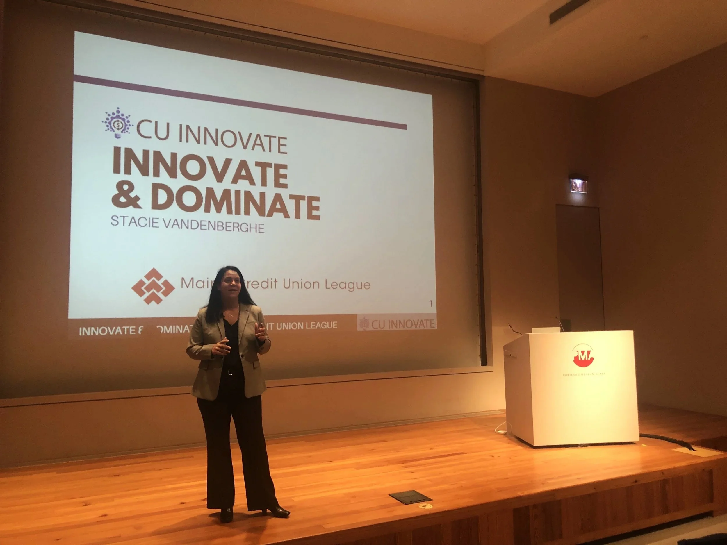 Woman presenting on a stage with a large screen behind her displaying a slide titled 'CU INNOVATE: Innovate & Dominate' by Stacie Vandenberghe, associated with Main Credit Union League, in a conference room with wooden flooring.