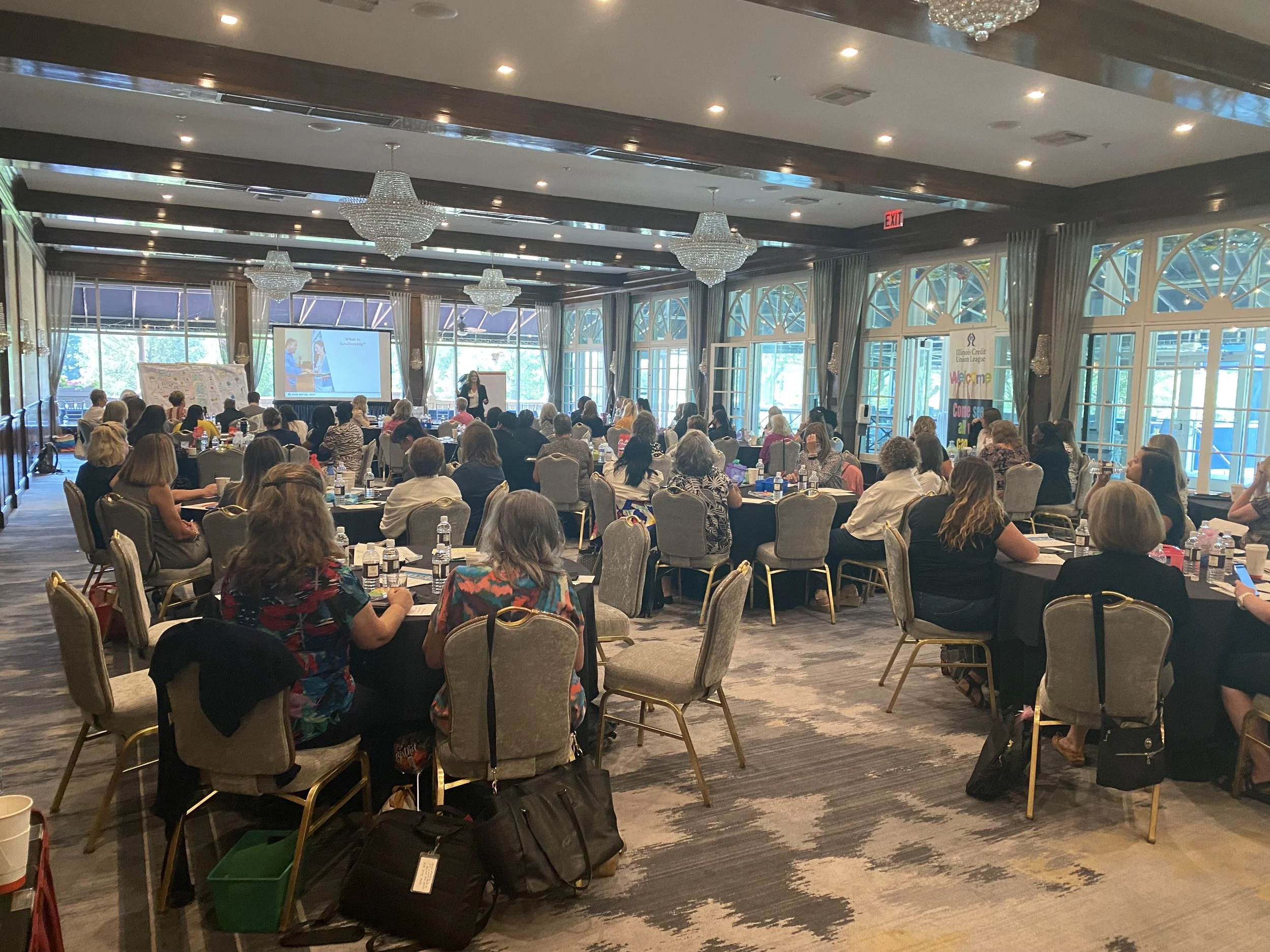 A large conference room filled with attendees seated at round tables, facing a stage with a speaker and a screen displaying a presentation, with large windows on the side.