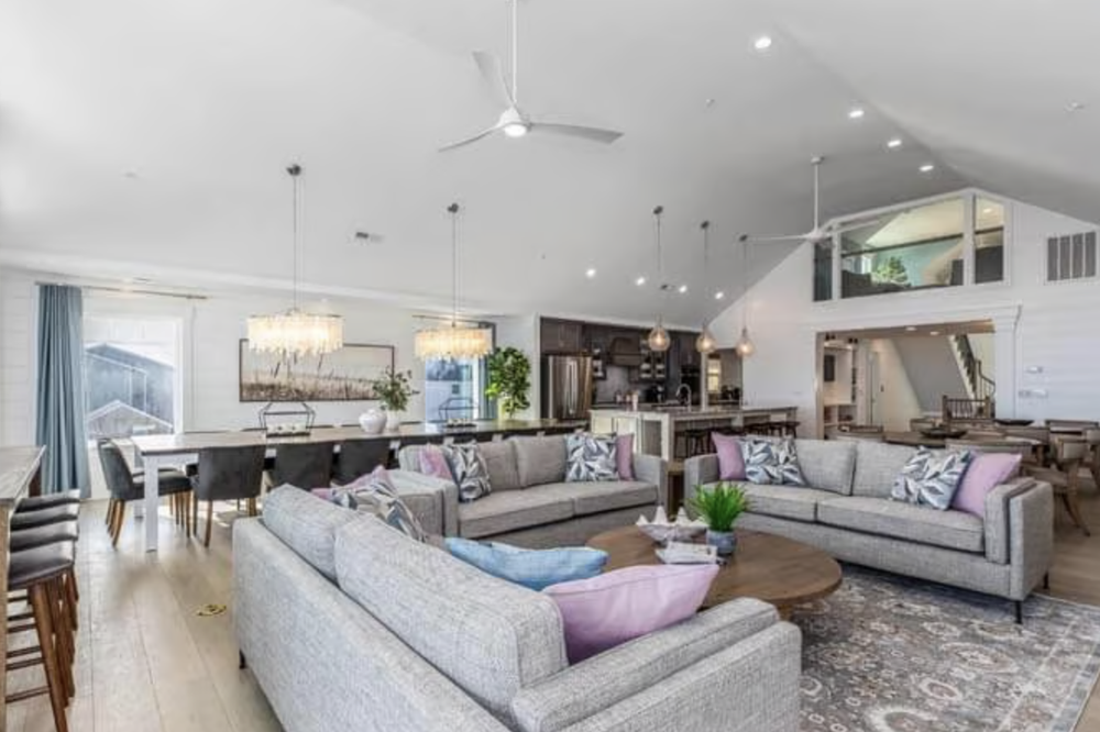 Bright open-concept living room with gray sofas, pink and patterned throw pillows, a wooden coffee table, and a large area rug. Dining area with a white table, black chairs, and three chandeliers. Kitchen with dark cabinets, stainless steel appliance