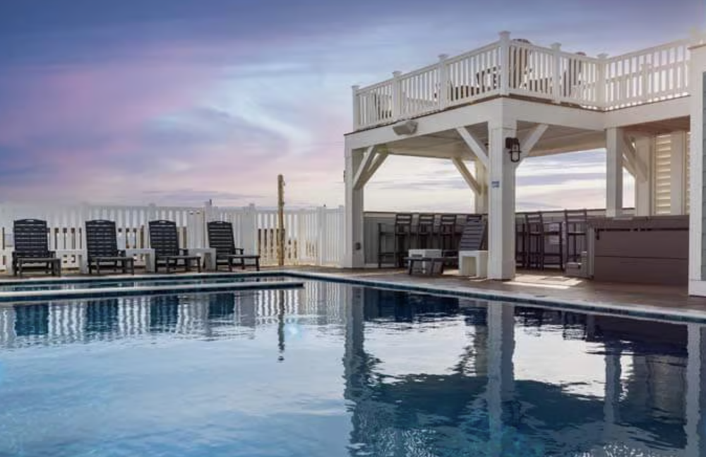 A backyard with a swimming pool, poolside chairs, and a white fenced area overlooking a colorful sunset sky.