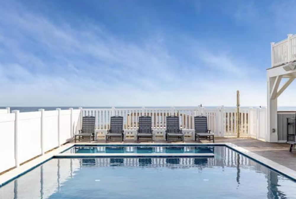Swimming pool with five lounge chairs on a deck, surrounded by a white fence, overlooking the ocean on a sunny day.