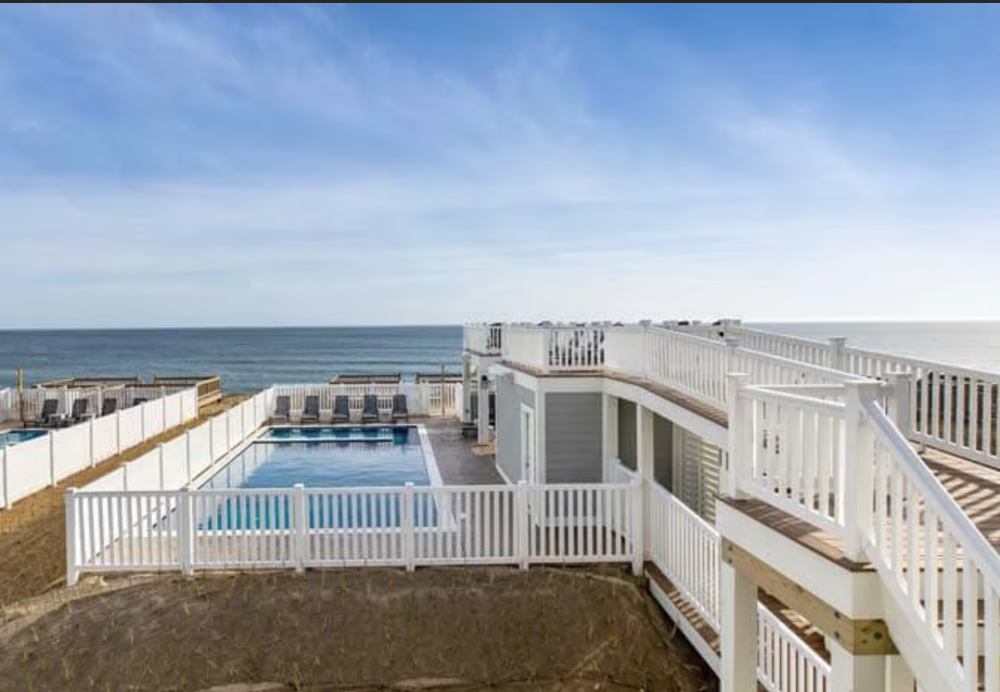 View of a beach house with a private pool, white fencing, and outdoor stairs, overlooking the ocean on a sunny day.