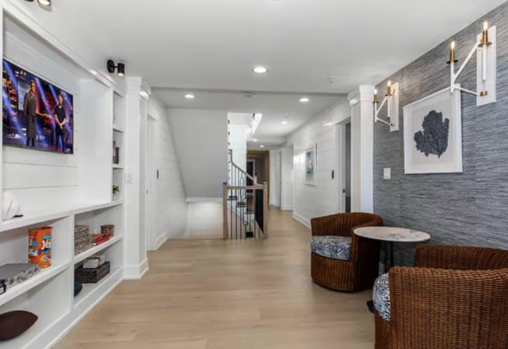 Interior of a modern living space with white walls, wooden flooring, a built-in bookshelf, wall-mounted TV, two brown wicker chairs with patterned cushions, a small round table, and decorative wall lighting.