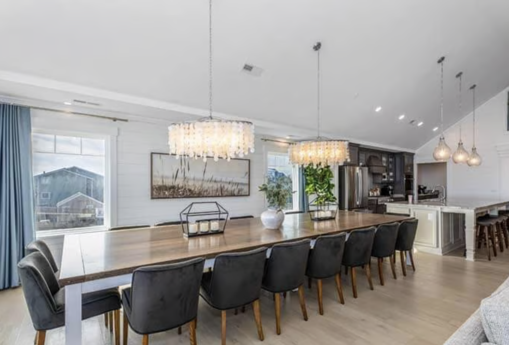 Modern dining room with a large wooden table, black chairs, and a chandelier. The room features white walls, large windows with blue curtains, and a view of neighboring houses. The kitchen area is visible in the background with stainless steel applia