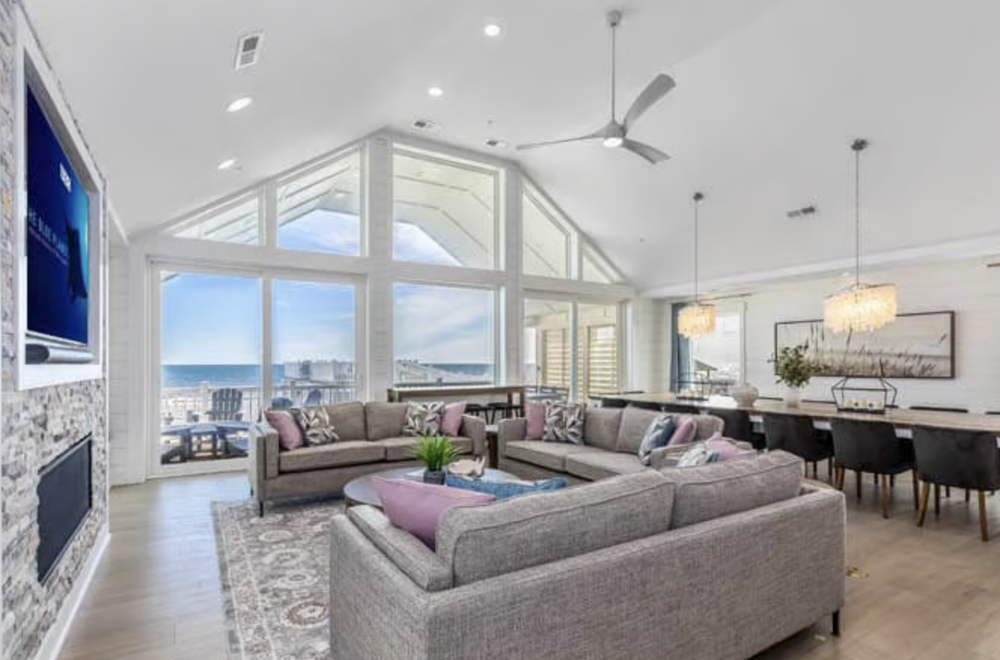 Living room with large windows offering ocean view, furnished with two gray sofas, colorful pillows, a coffee table, a fireplace, a TV, and a dining area with a wooden table and black chairs, decorated with pendant lights and artwork.