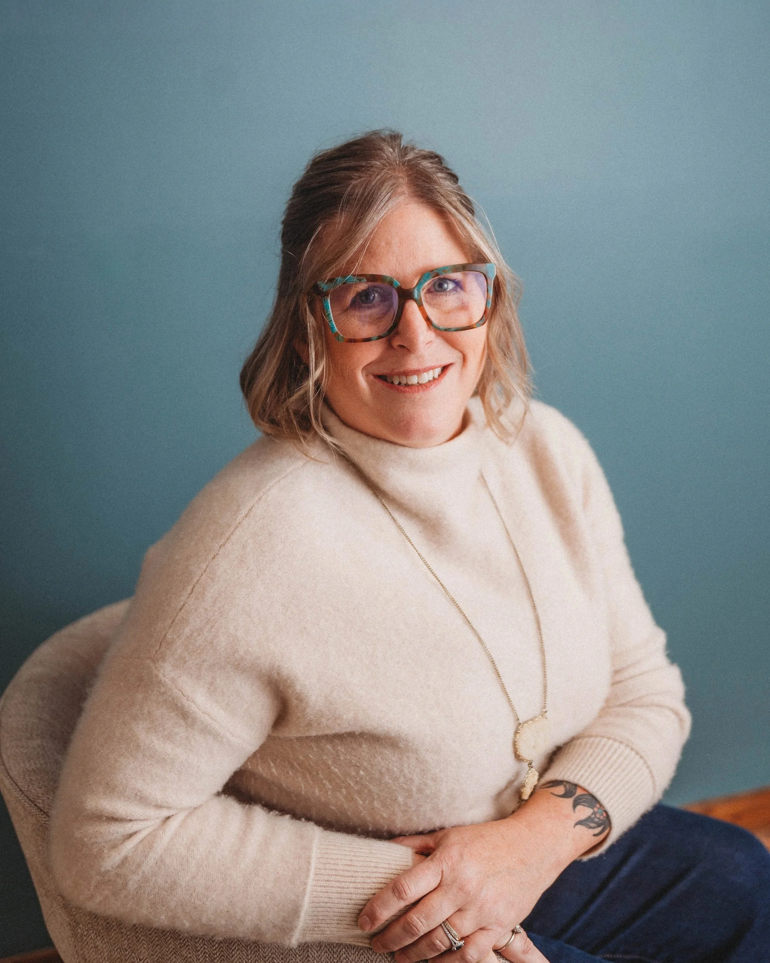 A woman with shoulder-length blonde hair, wearing large colorful glasses, a cream turtleneck sweater, and a long necklace, sitting against a teal wall.