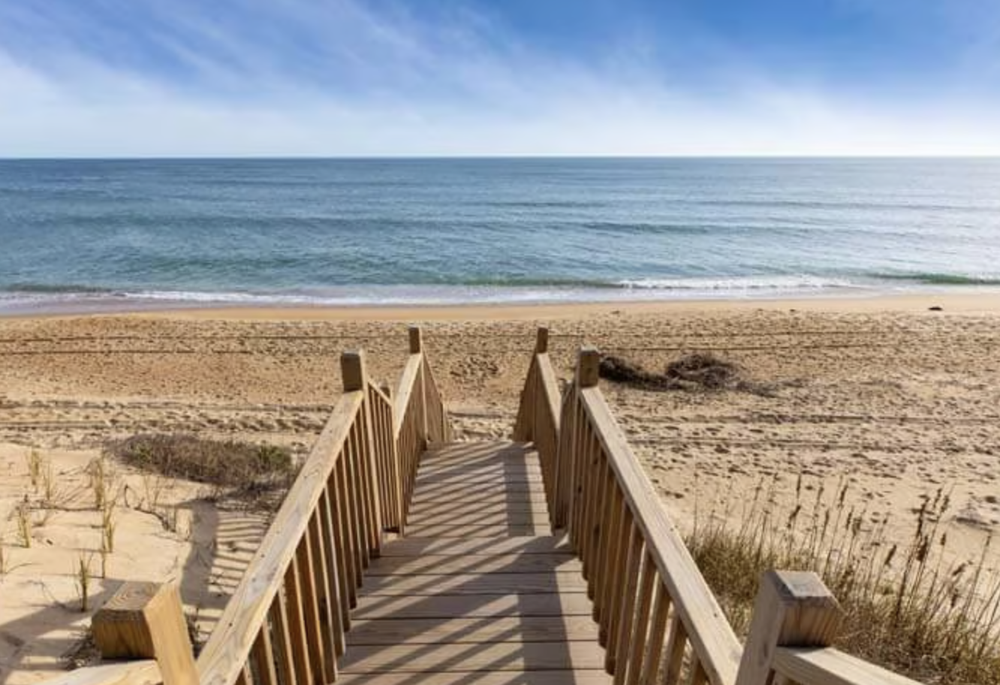 Wooden stairs leading down to a sandy beach with the ocean in the background and clear blue sky.