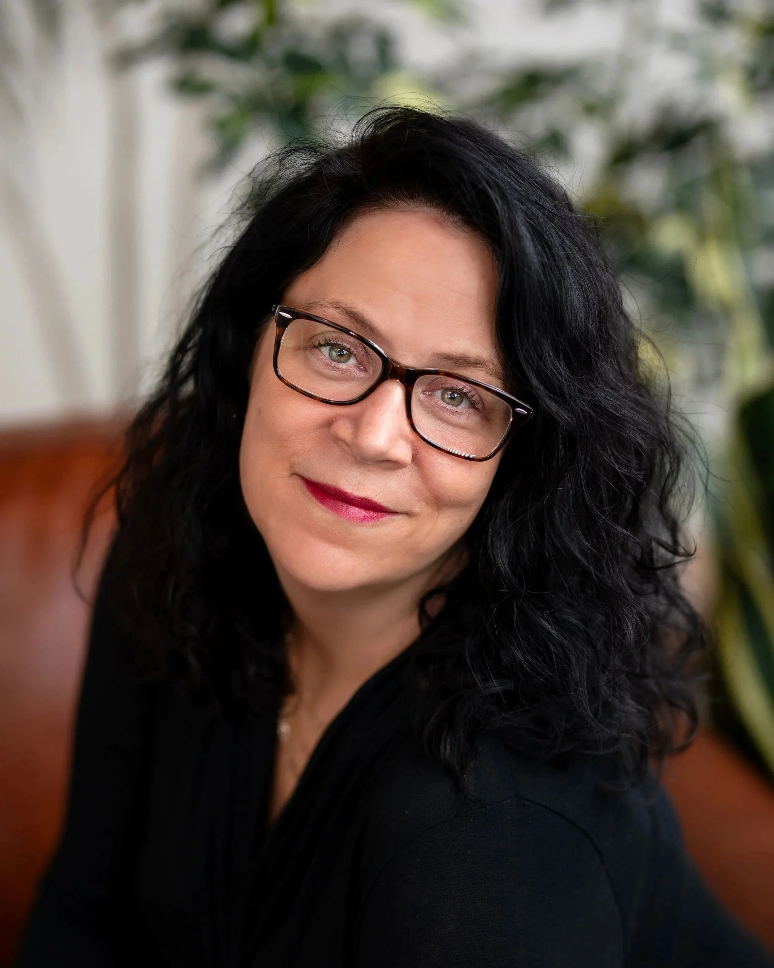 A woman with dark, curly hair and glasses smiling at the camera, sitting indoors with a blurred leafy background.
