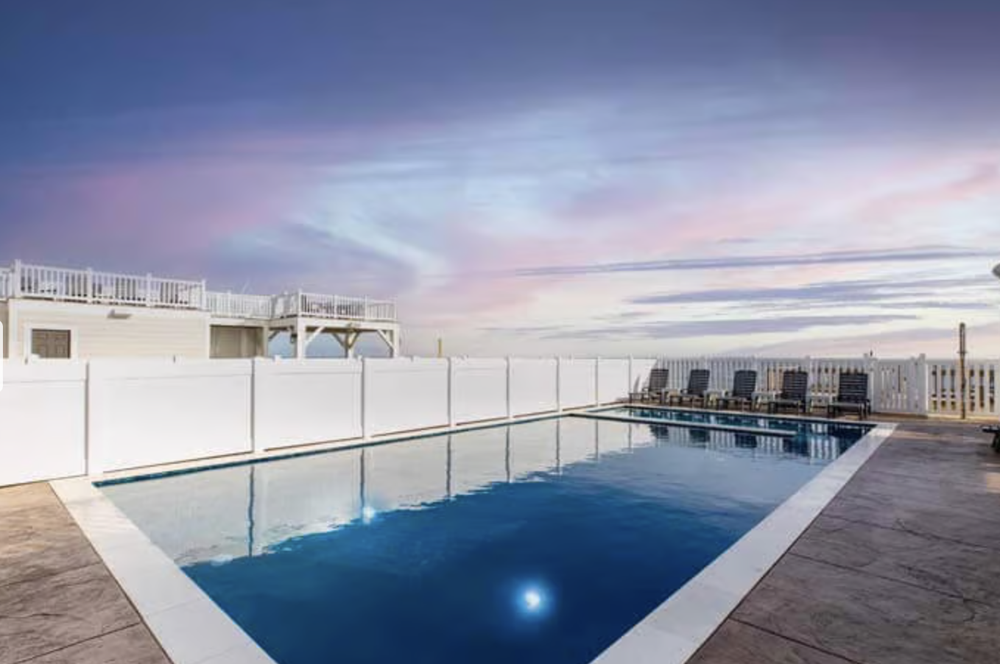 Empty swimming pool on a rooftop with lounge chairs and a white fence at sunset, pink and purple sky.