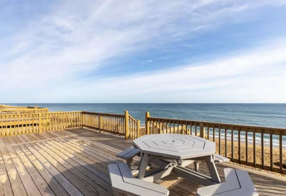 View of a wooden beach deck with a white picnic table, overlooking the ocean under a partly cloudy sky.