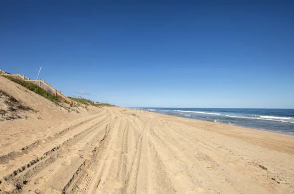 Empty sandy beach with tire tracks and ocean waves under a clear blue sky.