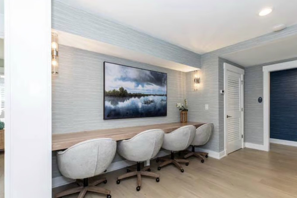 A room with a long wooden desk, four cream-colored chairs, a large landscape painting of a lake, and a small decorative plant on the desk. There are light gray textured walls, two wall sconces, and a closed white louvered door.