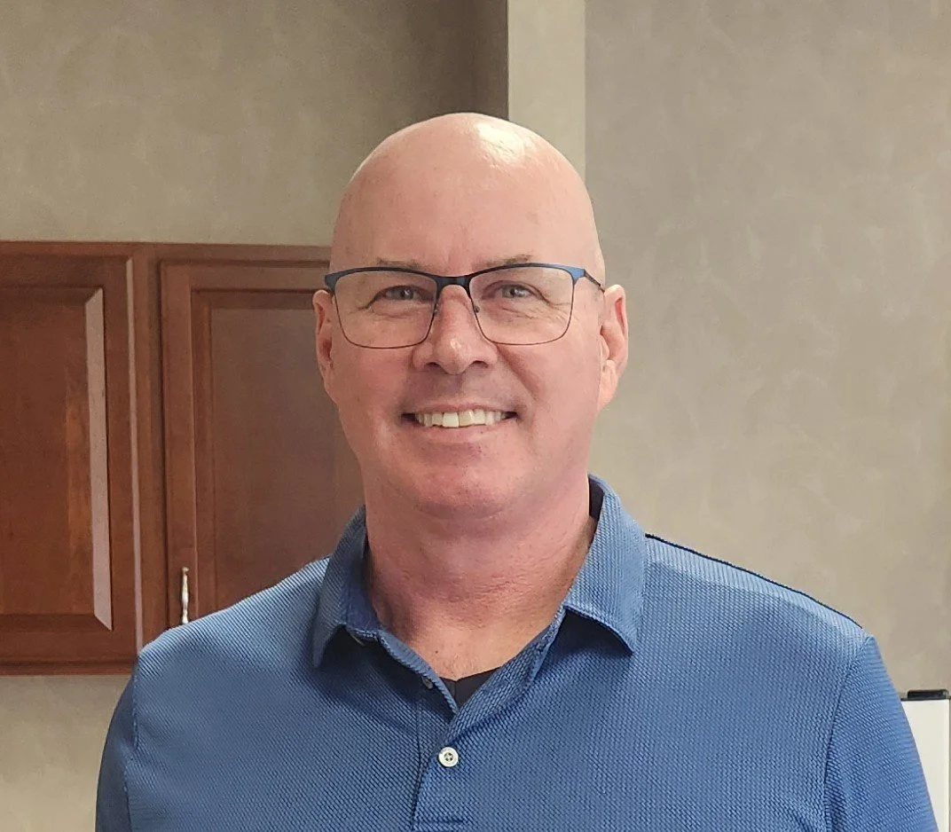 A smiling man with glasses and a blue collared shirt standing indoors.