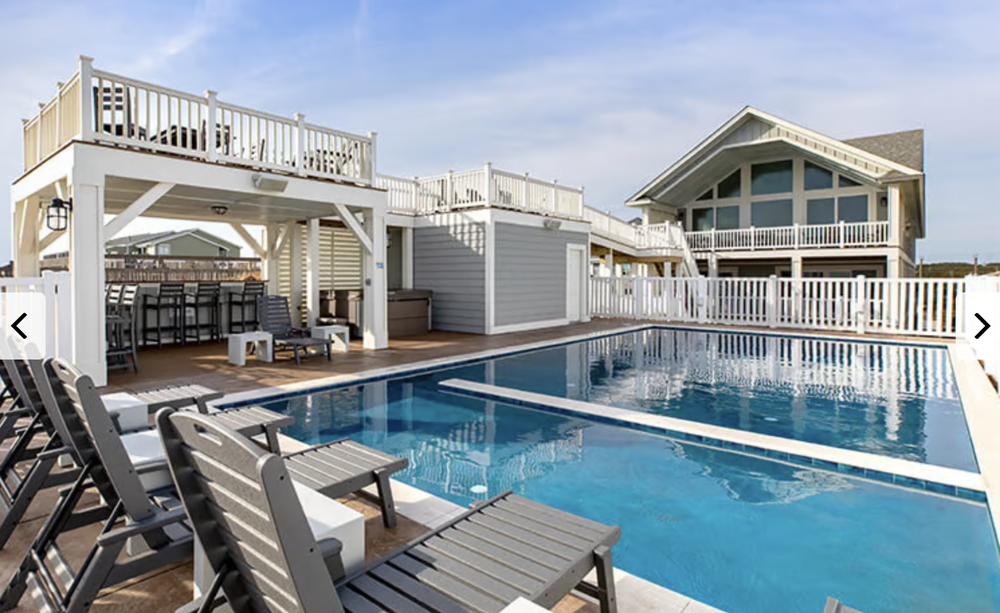 A rooftop swimming pool area with lounge chairs, a bar, and a multi-level deck overlooking other houses and a partly cloudy sky.