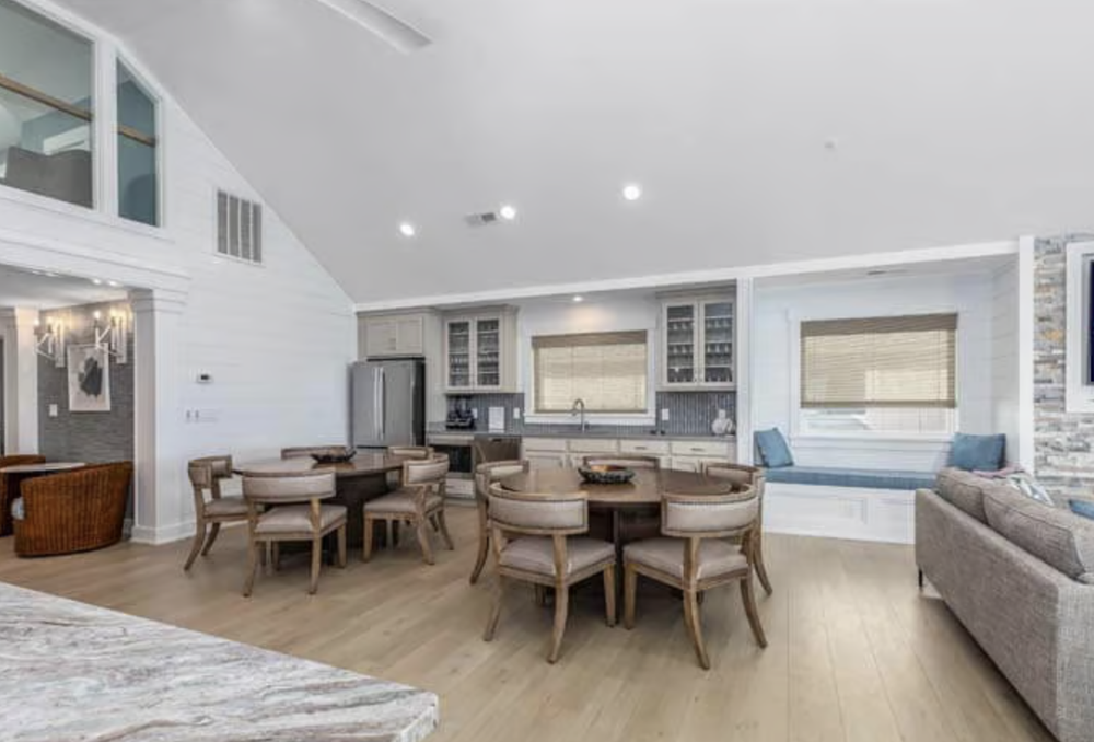 Open-concept living space with a kitchen, dining area, and sitting nook, featuring light wood flooring, white walls, and large windows with beige blinds.