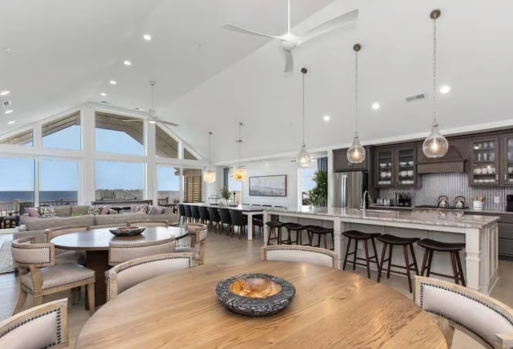 Spacious open-concept kitchen and living area with large windows showing an ocean view, wooden dining tables and chairs, bar-style stools at a kitchen island, modern lighting fixtures, vaulted ceiling, and ceiling fans.