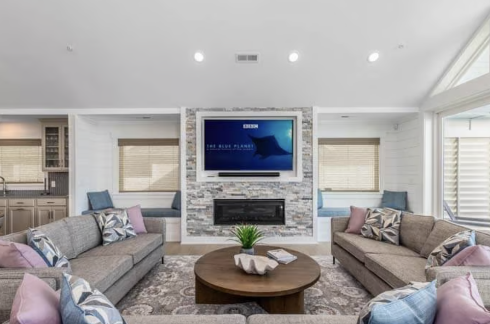 Living room with a stone fireplace, wall-mounted TV, beige couches with decorative pillows, round wooden coffee table, and built-in window seats with cushions and blinds.