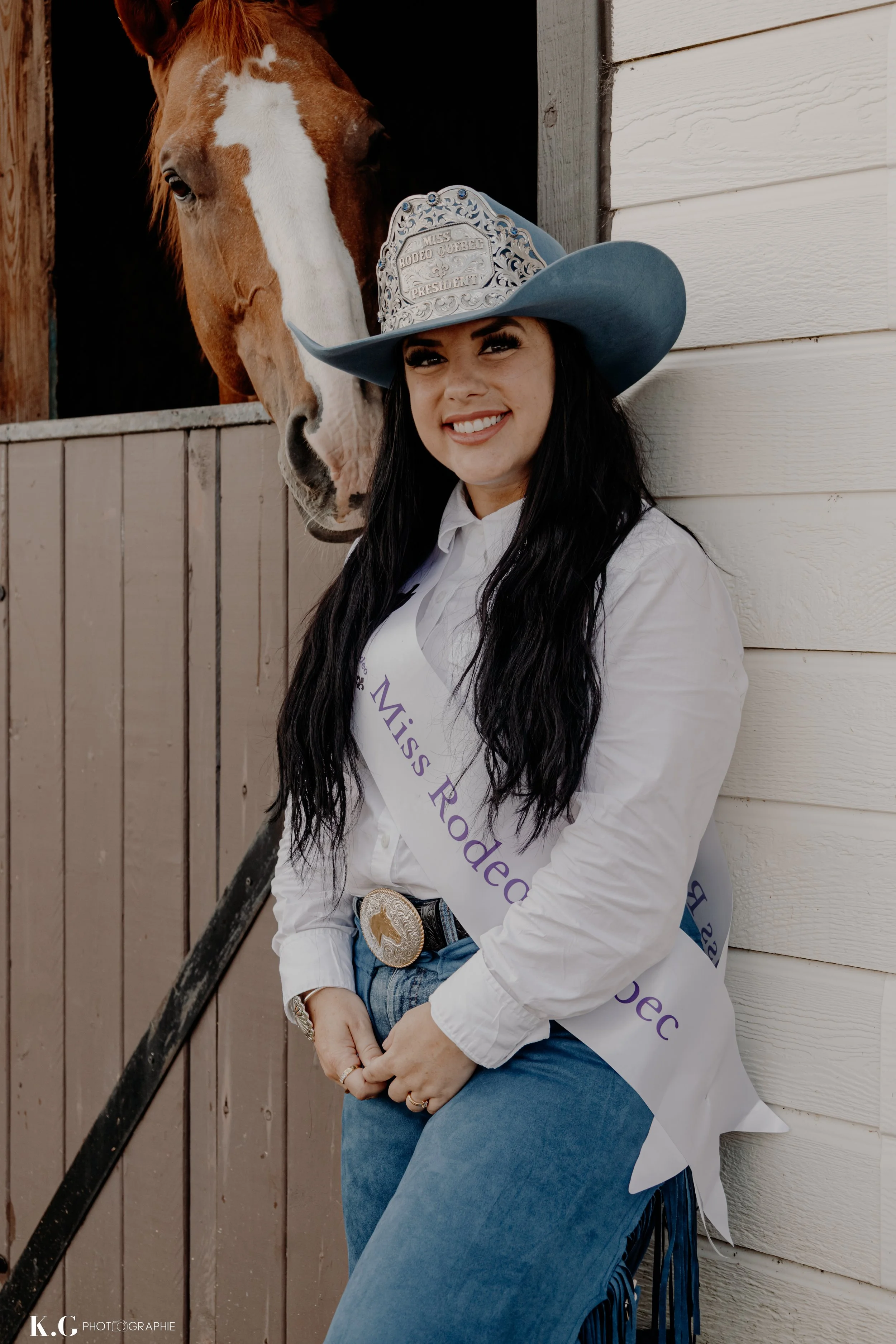 Jeune femme portant une couronne en tôle, une écharpe "Miss Rodeo Quebec 2023", une chemise blanche, un pantalon bleu, et une grande ceinture en argent, posant devant une stalle de cheval.