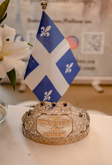 Une couronne en métal portant l'inscription "Miss Rodeo Québec Queen" et une petite drapeau du Québec avec une fleur de lys, accompagné de fleurs blanches à côté.