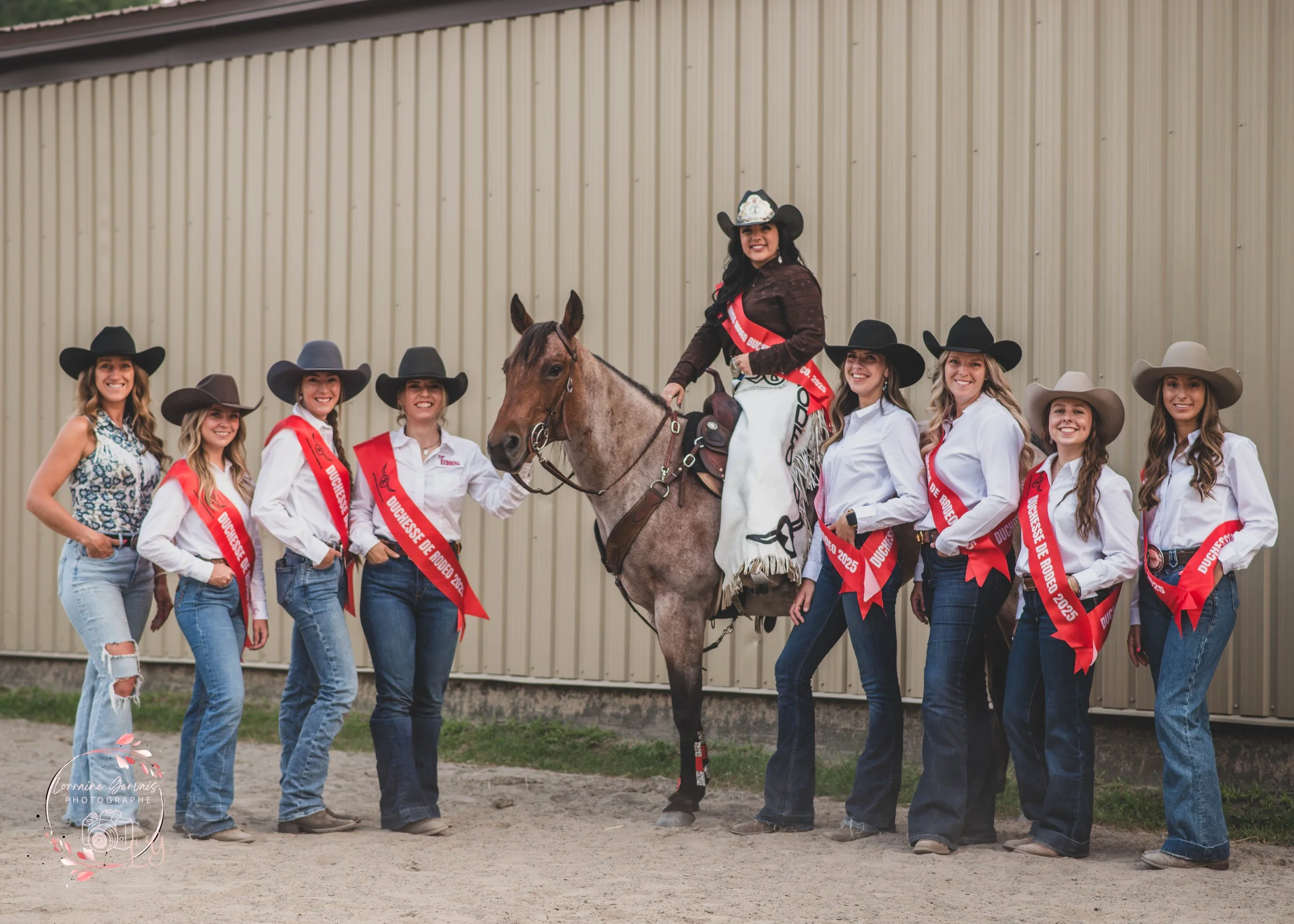 Groupe de femmes avec chapeaux et médailles, entourant une femme à cheval avec une sash, devant un mur en métal.