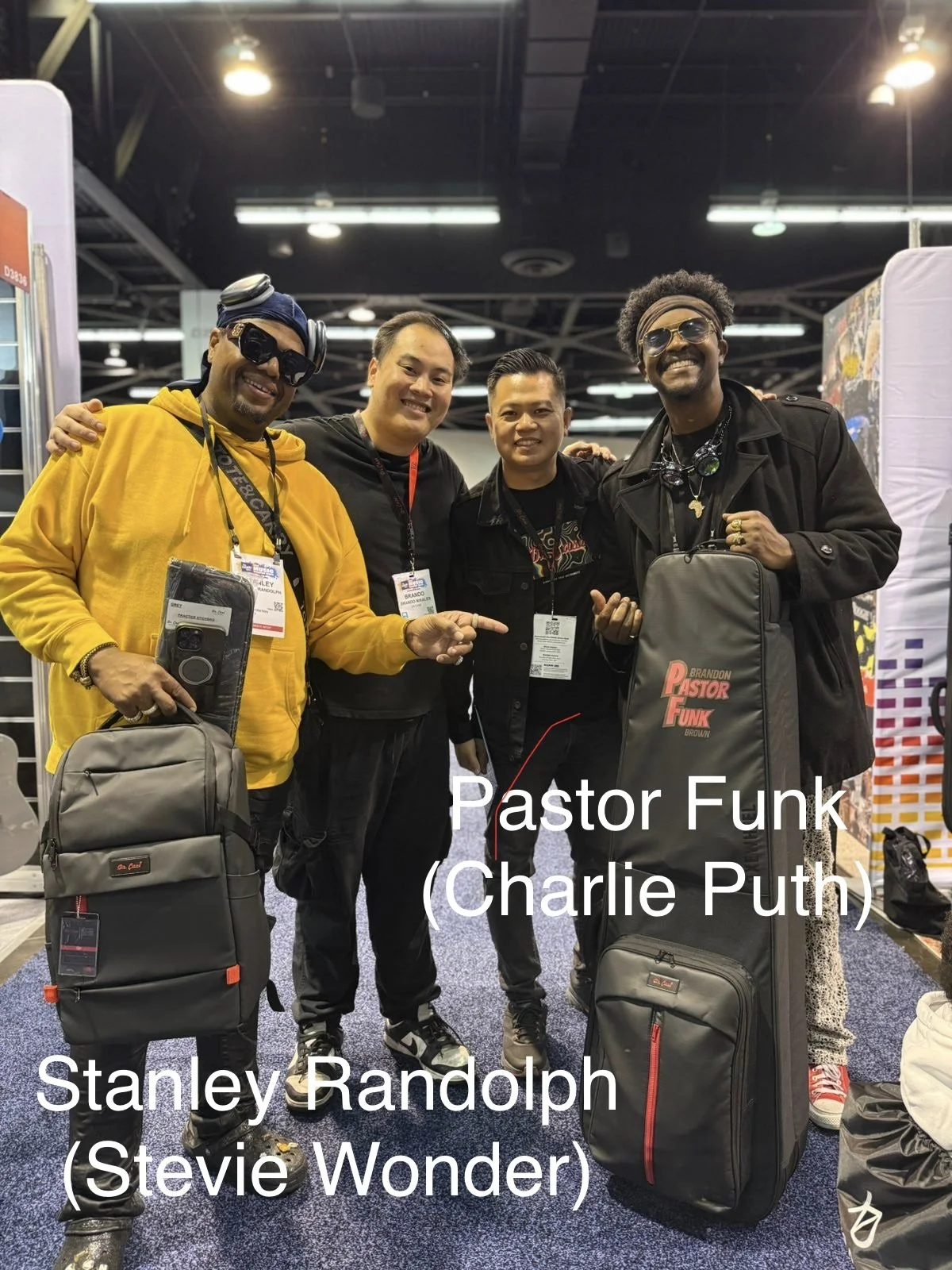 Four men standing together at an indoor event, smiling and holding personal items with luggage in front of and around them. Two men wear sunglasses and headphones, one man points at the camera, and all are wearing badges around their necks.