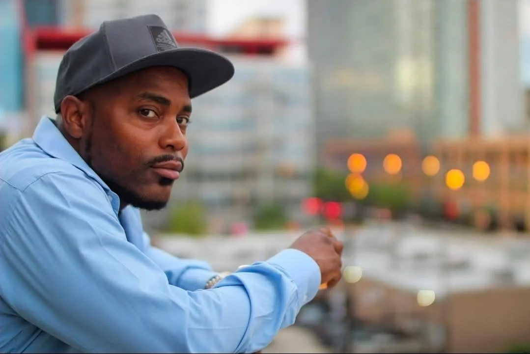 A man in a blue shirt and black cap sitting outdoors with a cityscape background at dusk.