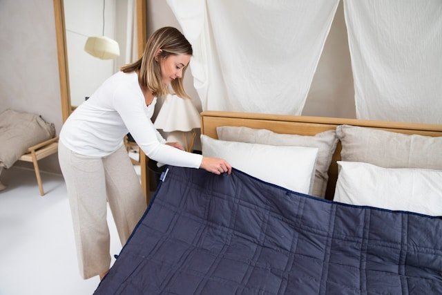 A woman making a bed with a navy blue quilted blanket in a bright bedroom.