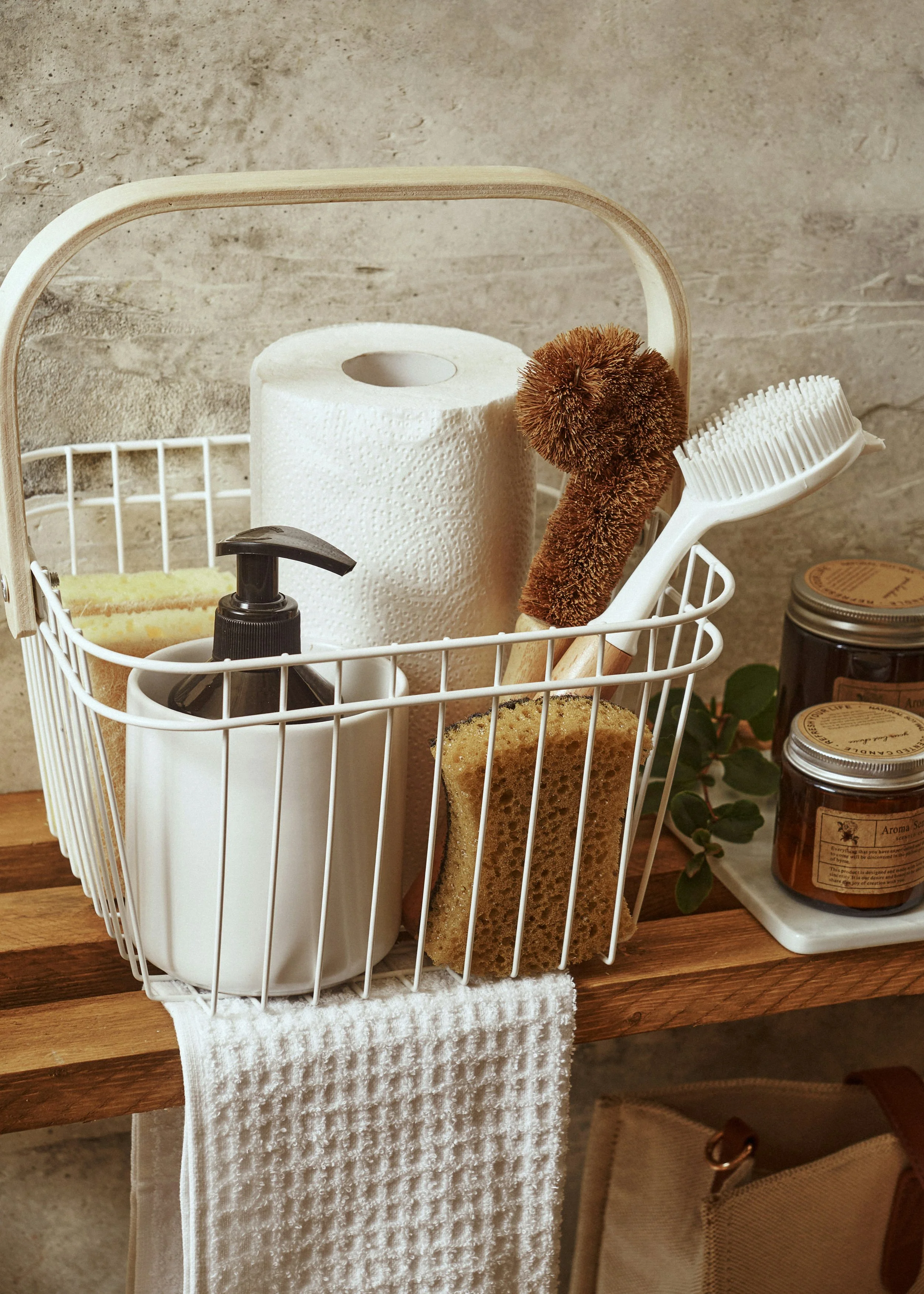 A white wire bathroom caddy containing a paper towel roll, a black soap dispenser, a sponge, a shower brush, and a brown loofah, placed on a wooden surface with jars of natural products nearby.