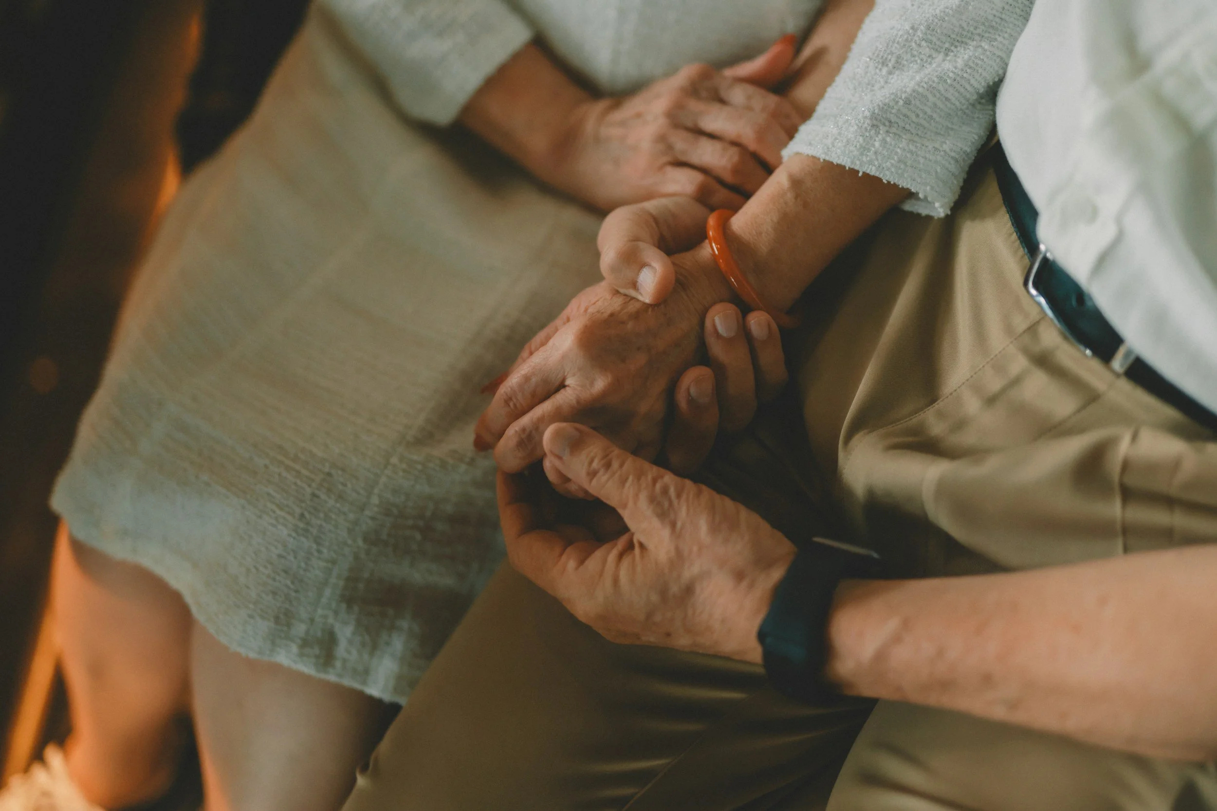 An elderly person with a red bracelet holding hands with a middle-aged person. The elderly person is sitting on a beige cushioned surface, wearing a light gray sweater, and has wrinkled skin and aged hands. The middle-aged person is standing, wearing khaki pants and a white shirt. The scene appears to be indoors, capturing a moment of support and connection.