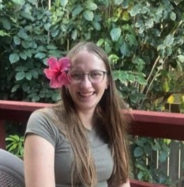Katy Schlaefer in front of green foliage and a reddish fence. Seated in a grey chair wearing a grey t-shirt with a pink hibiscus flower in Katy's long blonde hair. Katy has rounded wireframe glasses and is smiling to camera