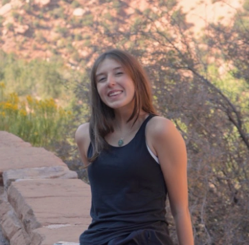 Jordyn Supernor sitting on a stone wall before a desert-like background with foliage. Jordyn is leaning back on both arms and is wearing a dark tank top with a necklace with a blue charm. Jordyn's lighter hair is longer than shoulders-length.