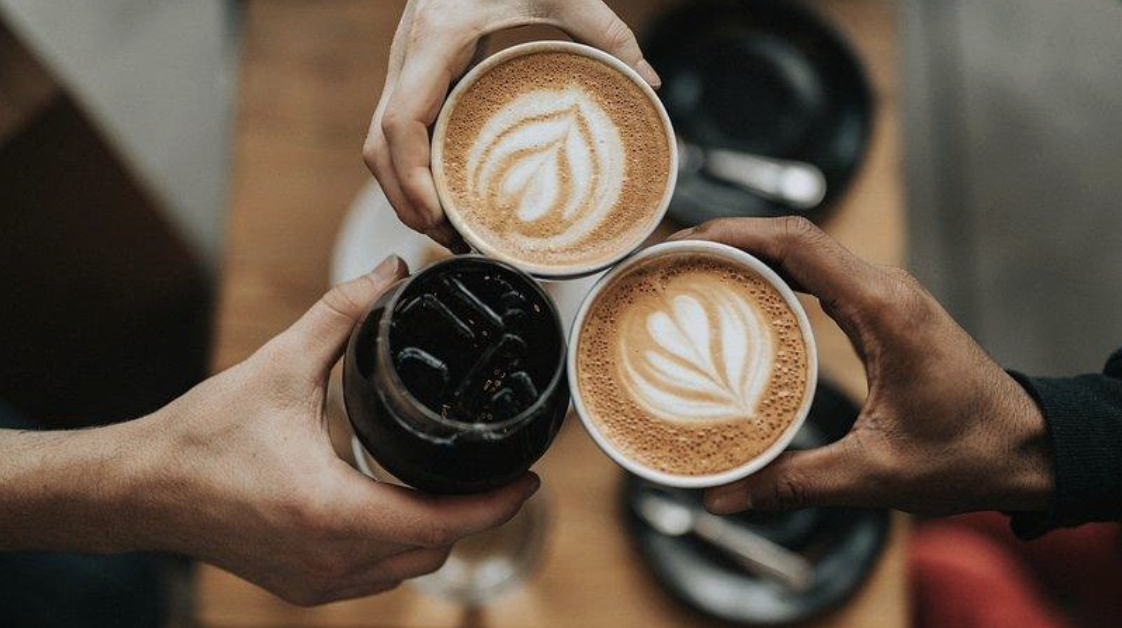 Three hands holding coffee together against a blurry background of a table. Two of the hands are holding hot coffees with latte art leaves and the third hand is holding a dark, black iced coffee.