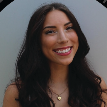 Lauren Saloio smiling to camera with a tilted head. Lauren has on a gold necklace with a heart charm and has long, curly black hair. Lauren has on a pinkish red lipstick