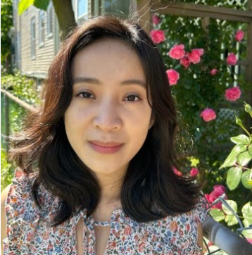 Vanan Phan standing in a garden with a house in the background behind pink flowers. Vanan is wearing a floral blouse. Vanan's hair is dark brown and just to the shoulders.
