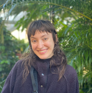 Lindsey Colby in front of green foliage. Lindsey is wearing a brownish jacket buttoned up with a black purse strap. Lindsey's brown hair is below shoulder-length and wavy. Lindsey is smiling to camera and has a nose piercing