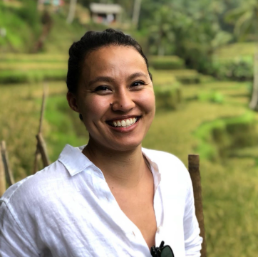 Veronique Lee in front of a green, hilly background, wearing a white linen button-up with dark sunglasses hanging from the shirt. Veronique's dark hair is pulled up and Veronique is smiling wide to camera